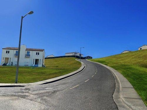A quiet road curving through grassy hills beside white houses under a blue sky.